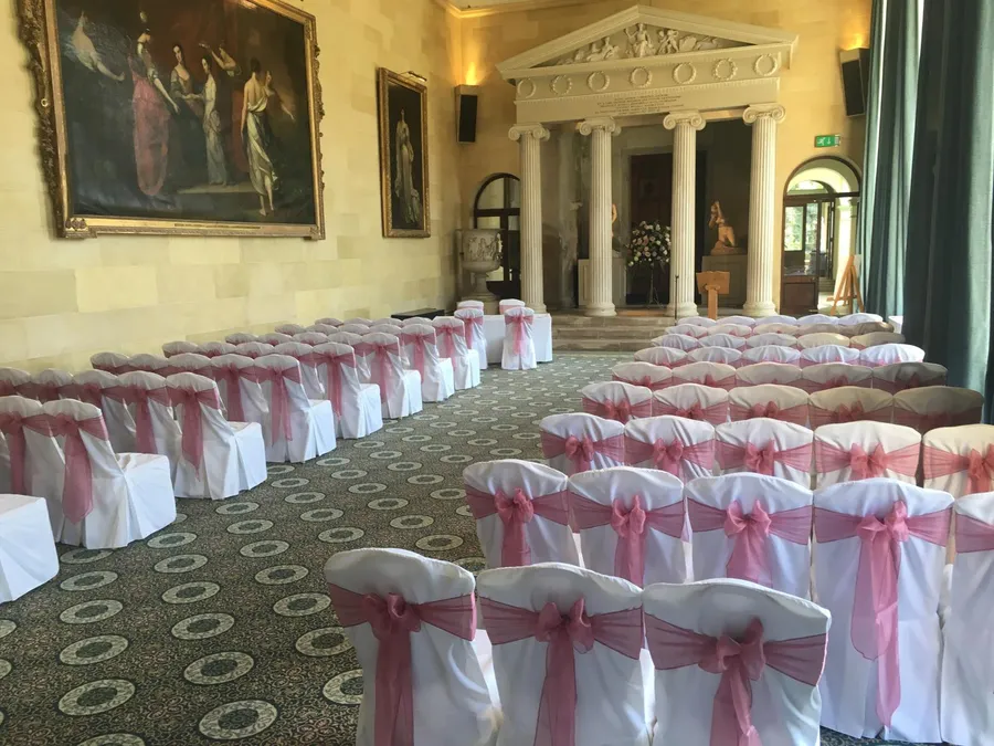 Rows of white chair covers with dusky-pink organza bows set out for a wedding ceremony in a grand gallery